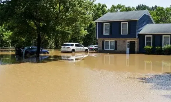 Brown floodwater surrounding a two-story house and parked cars after heavy storm rain